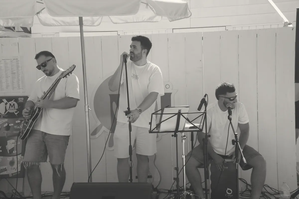 Playing guitar with two bandmates at an open-air bar, singer at the mic and cajón on the right.