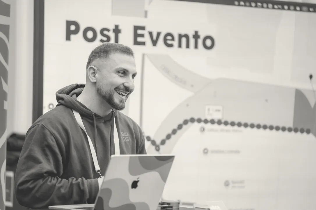 Laughing behind an open laptop, a slide titled Post Evento on the screen behind.