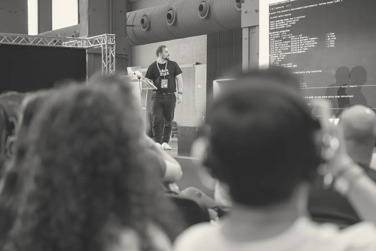 On a conference stage beside a lectern, a terminal session projected on the screen behind him.