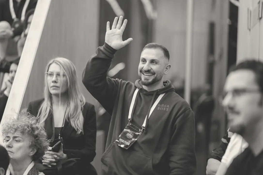 Standing in an audience with a visitor badge, waving with one hand raised.