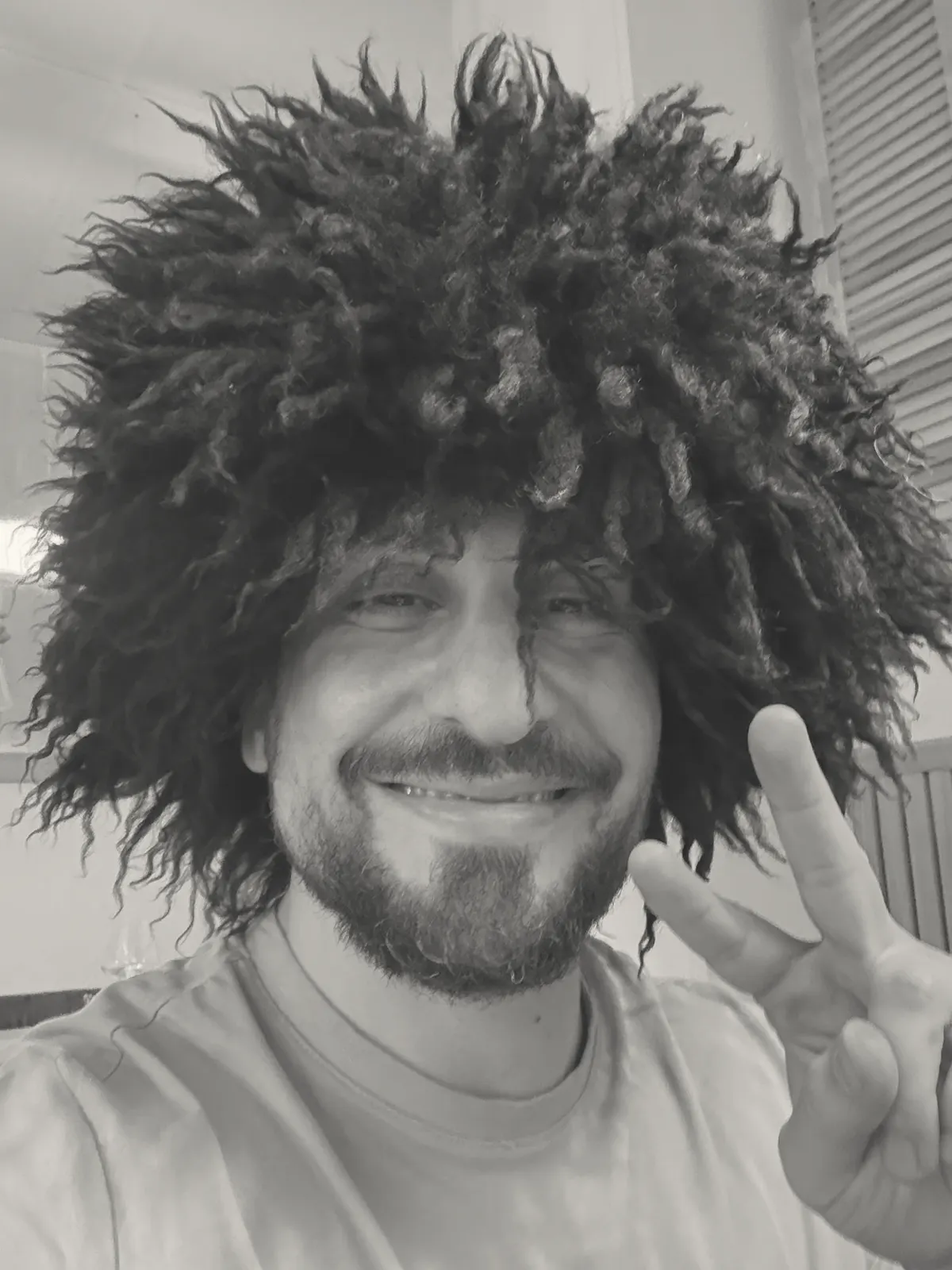 Grinning into a phone camera under an enormous curly wig, holding up two fingers.
